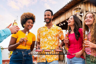 Multiethnic group of happy young friends having dinner barbecue party on rooftop at home - Multiracial cheerful young adult people having fun and bonding on a terrace balcony with city view, eating and drinking. - DMDF08561