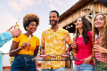 Multiethnische Gruppe von glücklichen jungen Freunden mit Abendessen Grillparty auf dem Dach zu Hause - Multirassische fröhliche junge erwachsene Menschen, die Spaß haben und sich auf einer Terrasse Balkon mit Blick auf die Stadt, Essen und Trinken. - DMDF08561