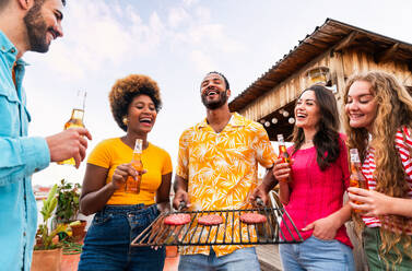 Multiethnische Gruppe von glücklichen jungen Freunden mit Abendessen Grillparty auf dem Dach zu Hause - Multirassische fröhliche junge erwachsene Menschen, die Spaß haben und sich auf einer Terrasse Balkon mit Blick auf die Stadt, Essen und Trinken. - DMDF08562