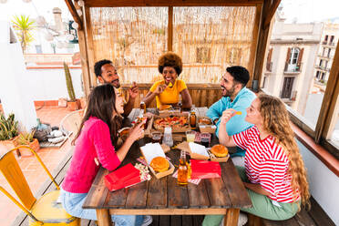 Multiethnic group of happy young friends having dinner barbecue party on rooftop at home - Multiracial cheerful young adult people having fun and bonding on a terrace balcony with city view, eating and drinking. - DMDF08573