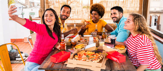 Multiethnic group of happy young friends having dinner barbecue party on rooftop at home - Multiracial cheerful young adult people having fun and bonding on a terrace balcony with city view, eating and drinking. - DMDF08578