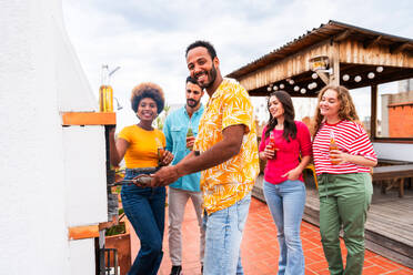 Multiethnische Gruppe von glücklichen jungen Freunden mit Abendessen Grillparty auf dem Dach zu Hause - Multirassische fröhliche junge erwachsene Menschen, die Spaß haben und sich auf einer Terrasse Balkon mit Blick auf die Stadt, Essen und Trinken. - DMDF08583