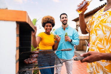 Multiethnic group of happy young friends having dinner barbecue party on rooftop at home - Multiracial cheerful young adult people having fun and bonding on a terrace balcony with city view, eating and drinking. - DMDF08585