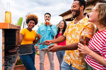 Multiethnische Gruppe von glücklichen jungen Freunden mit Abendessen Grillparty auf dem Dach zu Hause - Multirassische fröhliche junge erwachsene Menschen, die Spaß haben und sich auf einer Terrasse Balkon mit Blick auf die Stadt, Essen und Trinken. - DMDF08586