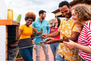 Multiethnische Gruppe von glücklichen jungen Freunden mit Abendessen Grillparty auf dem Dach zu Hause - Multirassische fröhliche junge erwachsene Menschen, die Spaß haben und sich auf einer Terrasse Balkon mit Blick auf die Stadt, Essen und Trinken. - DMDF08587