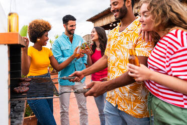 Multiethnic group of happy young friends having dinner barbecue party on rooftop at home - Multiracial cheerful young adult people having fun and bonding on a terrace balcony with city view, eating and drinking. - DMDF08588