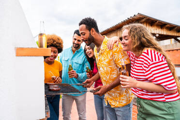 Multiethnische Gruppe von glücklichen jungen Freunden mit Abendessen Grillparty auf dem Dach zu Hause - Multirassische fröhliche junge erwachsene Menschen, die Spaß haben und sich auf einer Terrasse Balkon mit Blick auf die Stadt, Essen und Trinken. - DMDF08589