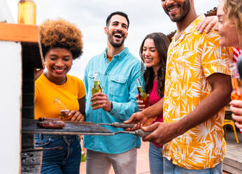 Multiethnic group of happy young friends having dinner barbecue party on rooftop at home - Multiracial cheerful young adult people having fun and bonding on a terrace balcony with city view, eating and drinking. - DMDF08590