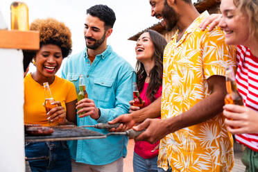 Multiethnic group of happy young friends having dinner barbecue party on rooftop at home - Multiracial cheerful young adult people having fun and bonding on a terrace balcony with city view, eating and drinking. - DMDF08592