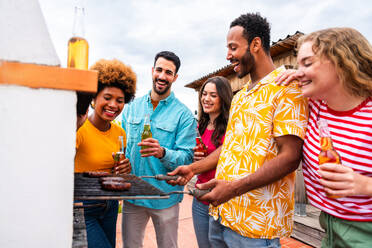 Multiethnische Gruppe von glücklichen jungen Freunden mit Abendessen Grillparty auf dem Dach zu Hause - Multirassische fröhliche junge erwachsene Menschen, die Spaß haben und sich auf einer Terrasse Balkon mit Blick auf die Stadt, Essen und Trinken. - DMDF08593