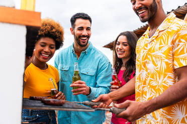 Multiethnic group of happy young friends having dinner barbecue party on rooftop at home - Multiracial cheerful young adult people having fun and bonding on a terrace balcony with city view, eating and drinking. - DMDF08594