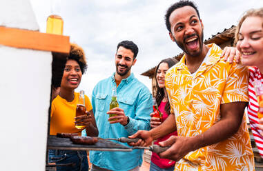 Multiethnische Gruppe von glücklichen jungen Freunden mit Abendessen Grillparty auf dem Dach zu Hause - Multirassische fröhliche junge erwachsene Menschen, die Spaß haben und sich auf einer Terrasse Balkon mit Blick auf die Stadt, Essen und Trinken. - DMDF08595