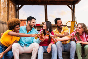 Multiethnic group of happy young friends having dinner barbecue party on rooftop at home - Multiracial cheerful young adult people having fun and bonding on a terrace balcony with city view, eating and drinking. - DMDF08601