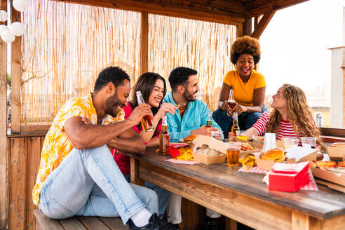 Multiethnische Gruppe von glücklichen jungen Freunden mit Abendessen Grillparty auf dem Dach zu Hause - Multirassische fröhliche junge erwachsene Menschen, die Spaß haben und sich auf einer Terrasse Balkon mit Blick auf die Stadt, Essen und Trinken. - DMDF08604