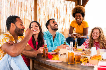 Multiethnic group of happy young friends having dinner barbecue party on rooftop at home - Multiracial cheerful young adult people having fun and bonding on a terrace balcony with city view, eating and drinking. - DMDF08608