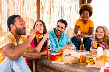 Multiethnic group of happy young friends having dinner barbecue party on rooftop at home - Multiracial cheerful young adult people having fun and bonding on a terrace balcony with city view, eating and drinking. - DMDF08609