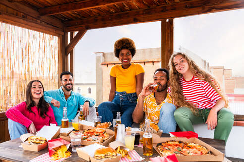 Multiethnische Gruppe von glücklichen jungen Freunden mit Abendessen Grillparty auf dem Dach zu Hause - Multirassische fröhliche junge erwachsene Menschen, die Spaß haben und sich auf einer Terrasse Balkon mit Blick auf die Stadt, Essen und Trinken. - DMDF08612