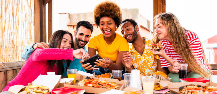 Multiethnische Gruppe von glücklichen jungen Freunden mit Abendessen Grillparty auf dem Dach zu Hause - Multirassische fröhliche junge erwachsene Menschen, die Spaß haben und sich auf einer Terrasse Balkon mit Blick auf die Stadt, Essen und Trinken. - DMDF08621