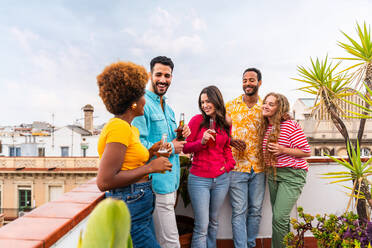 Multiethnic group of happy young friends having dinner barbecue party on rooftop at home - Multiracial cheerful young adult people having fun and bonding on a terrace balcony with city view, eating and drinking. - DMDF08648