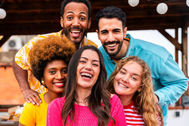 Multiethnic group of happy young friends having dinner barbecue party on rooftop at home - Multiracial cheerful young adult people having fun and bonding on a terrace balcony with city view, eating and drinking. Close-up portrait of funny faces. - DMDF08657