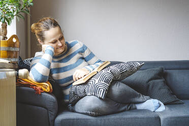 Smiling woman reading book on sofa at home - IHF01860