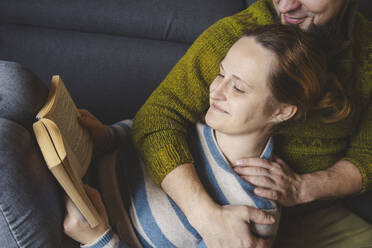 Smiling woman lying on man's lap and reading book at home - IHF01863