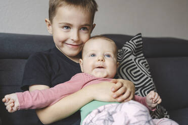 Smiling boy holding baby sister on sofa at home - IHF01866