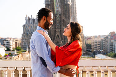 Multiracial beautiful happy couple of lovers dating on rooftop balcony at Sagrada Familia, Barcelona - Multiethnic people having romantic meeting on a terrace with city view , concepts about tourism and people lifestyle - DMDF08779