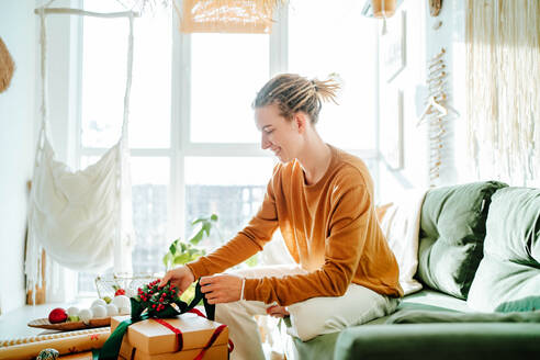 Side view of Young blond hair woman in casual clothes smiling and preparing wrapped gift box while sitting on comfortable sofa at home - ADSF51008