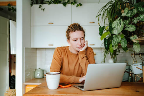 Thoughtful young female with dreadlocks looking at screen of laptop and reading recipe while sitting leaning chin on hand at kitchen table with mobile phone and mug against cabinets and green plants - ADSF51014