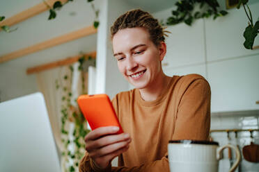Low angle of Smiling young female in casual clothes and with dreadlocks looking at screen of mobile phone while sitting at kitchen table with laptop coffee mug and with toothy smile reading text messages near plants - ADSF51016