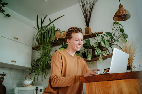Low angle of Smiling young female with dreadlocks looking at screen of laptop while while working at kitchen table with mobile phone and mug against cabinets and green plants - ADSF51020