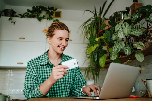 Young female with dreadlocks looking at screen of laptop while sitting with credit card in hand and with toothy smile doing online purchases in room with green plants in daylight - ADSF51024