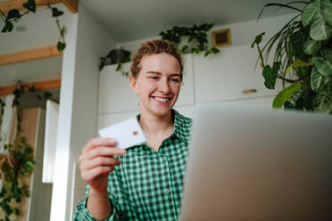 Low angle of smiling young female with dreadlocks looking at screen of laptop while sitting with credit card in hand and with toothy smile doing online purchases in room with green plants in daylight - ADSF51025