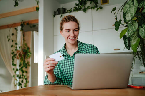 Young female with dreadlocks looking at screen of laptop while sitting with credit card in hand and with toothy smile doing online purchases in room with green plants in daylight - ADSF51026