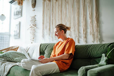 Side view of Young female freelancer smiling and working on project while sitting on comfortable sofa with cushions at home near window - ADSF51033