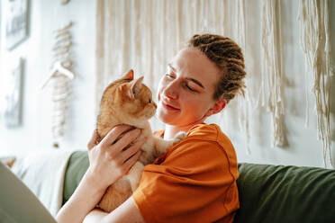 Young woman embracing adorable British shorthair golden cat while sitting on comfortable couch against blurred interior of living room at home - ADSF51035