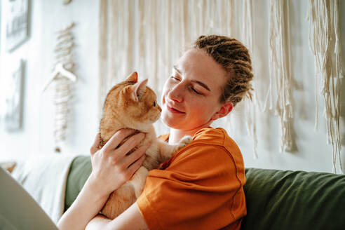 Young woman embracing adorable British shorthair golden cat while sitting on comfortable couch against blurred interior of living room at home - ADSF51035