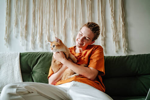 Young woman embracing adorable British shorthair golden cat while sitting on comfortable couch against blurred interior of living room at home - ADSF51036