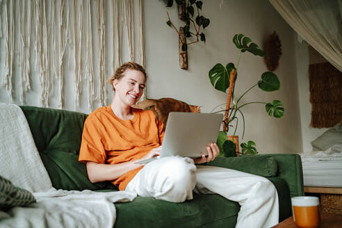 Young female freelancer smiling and working on project while sitting on comfortable sofa with cushions at home near adorable British shorthair golden cat - ADSF51037