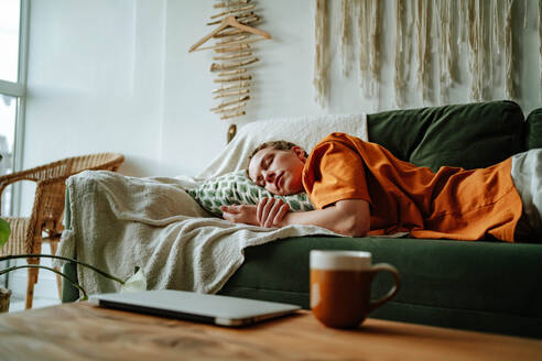 Young woman sleeping on comfortable sofa with cushion at home near laptop and cup on table in cozy living room of modern apartment - ADSF51040