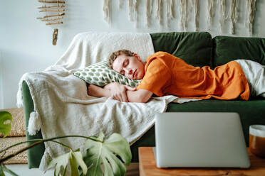 Young woman sleeping on comfortable sofa with cushion at home near laptop and cup on table in cozy living room of modern apartment - ADSF51042