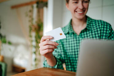 Closeup of blurred smiling young female looking at screen of laptop while sitting with credit card in hand and with toothy smile doing online purchases in room in daylight - ADSF51043