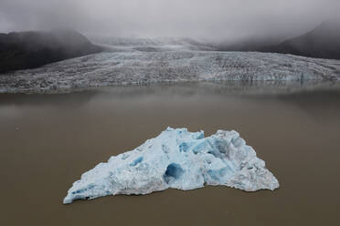 A solitary iceberg floats in a serene glacial lagoon against a backdrop of a vast glacier under moody skies in Iceland - ADSF51084