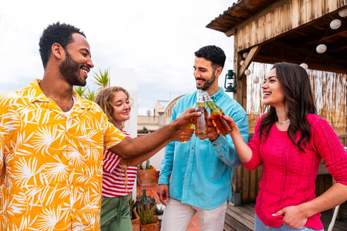 Multiethnic group of happy young friends having dinner barbecue party on rooftop at home - Multiracial cheerful young adult people having fun and bonding on a terrace balcony with city view, eating and drinking. - DMDF09166