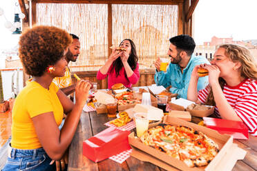 Multiethnic group of happy young friends having dinner barbecue party on rooftop at home - Multiracial cheerful young adult people having fun and bonding on a terrace balcony with city view, eating and drinking. - DMDF09178