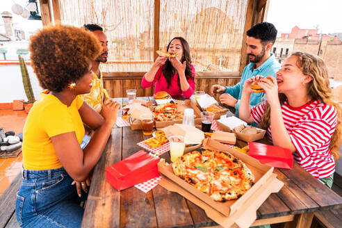 Multiethnic group of happy young friends having dinner barbecue party on rooftop at home - Multiracial cheerful young adult people having fun and bonding on a terrace balcony with city view, eating and drinking. - DMDF09179