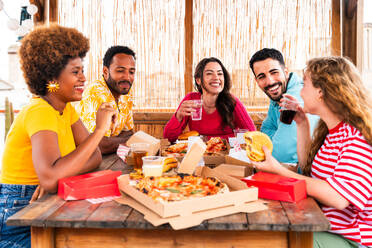 Multiethnic group of happy young friends having dinner barbecue party on rooftop at home - Multiracial cheerful young adult people having fun and bonding on a terrace balcony with city view, eating and drinking. - DMDF09181