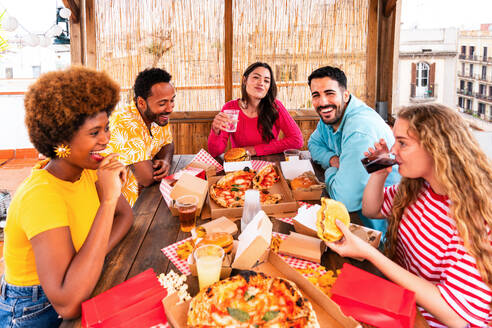 Multiethnic group of happy young friends having dinner barbecue party on rooftop at home - Multiracial cheerful young adult people having fun and bonding on a terrace balcony with city view, eating and drinking. - DMDF09182