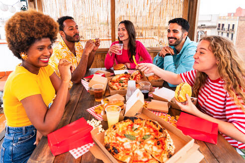 Multiethnic group of happy young friends having dinner barbecue party on rooftop at home - Multiracial cheerful young adult people having fun and bonding on a terrace balcony with city view, eating and drinking. - DMDF09183